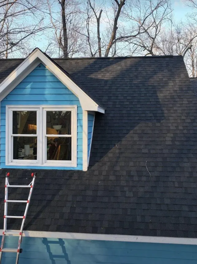 An aerial view of a house shows a newly installed dark shingle roof with a light blue dormer featuring a white framed window. A silver ladder with red caps rests on the roof near the dormer.